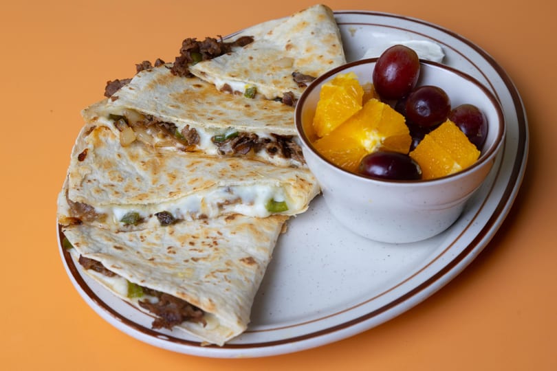 Cheesesteak quesadillas with melted cheese and green peppers, served with a side of fruit.