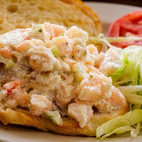 Shrimp salad on toasted bread with shredded lettuce and sliced tomatoes on a white plate.