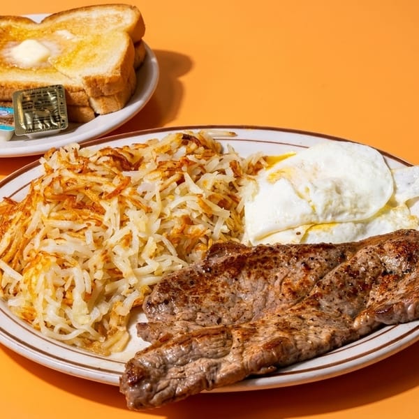 Steak and eggs breakfast with hash browns and buttered toast on an orange background.